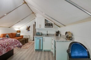 Habitación compacta y bien iluminada con kitchenette, una cama con ropa de cama estampada y una silla azul.