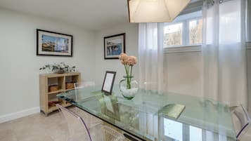 A dining area with a glass table, clear chairs, a vase with flowers, and two framed pictures on the wall.