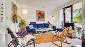 Una sala de estar acogedora con un sofá azul, un sillón de mimbre, una mecedora y una mesa de centro de vidrio. Hay un cuadro en la pared y una planta en maceta.