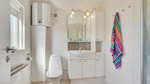 Un baño pequeño con inodoro, lavabo y espejo. Hay un perchero con una toalla de colores, y una ventana con persianas.
