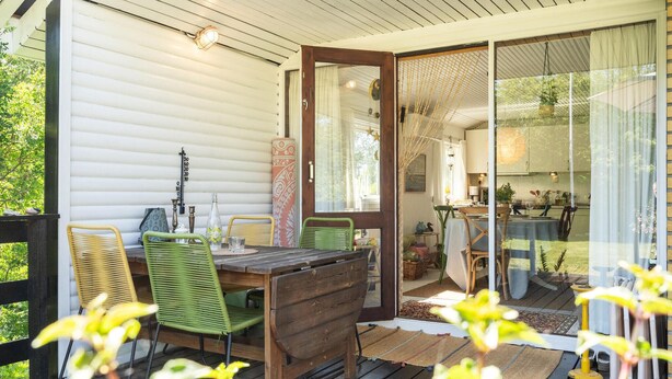 Un área de comedor exterior acogedora con una mesa y sillas de madera, un vidrio que da a la cocina y una terraza de madera.