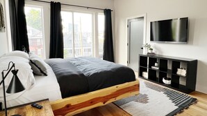 A modern bedroom with a wooden bed frame, a flat-screen TV, a bedside table, and a view of the city through large windows. - New! Downtown, Rooftop, Walkable to The Gulch, Music Row & 1M to Broadway! (Nashville)