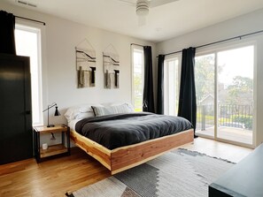 Une chambre moderne avec un lit en bois, une table de chevet, une lampe et une vue sur l’extérieur grâce à de grandes fenêtres.