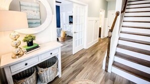 A hallway with a wooden floor, a white console table with a mirror, and a staircase with wooden steps. - A Block from the Dock in Beau Coast Classic (Beaufort)