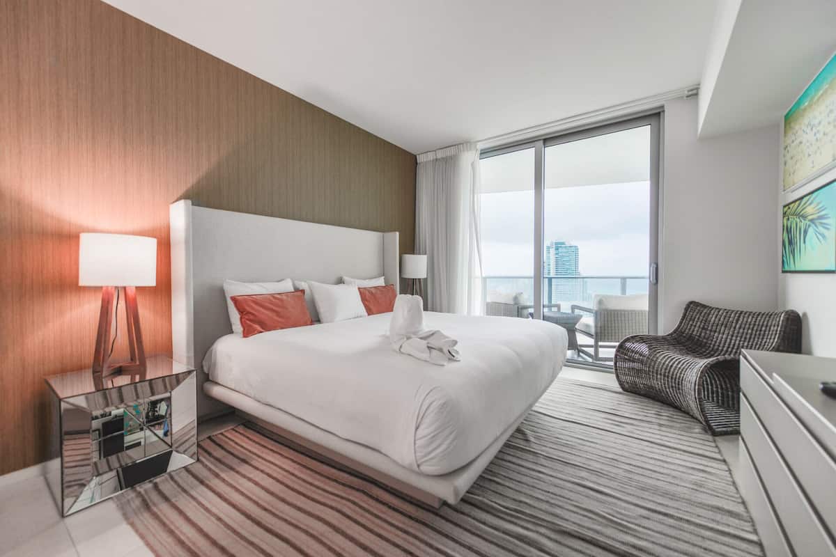 Une chambre d’hôtel avec un grand lit, une vue sur la ville, une lampe moderne et un fauteuil en osier.
