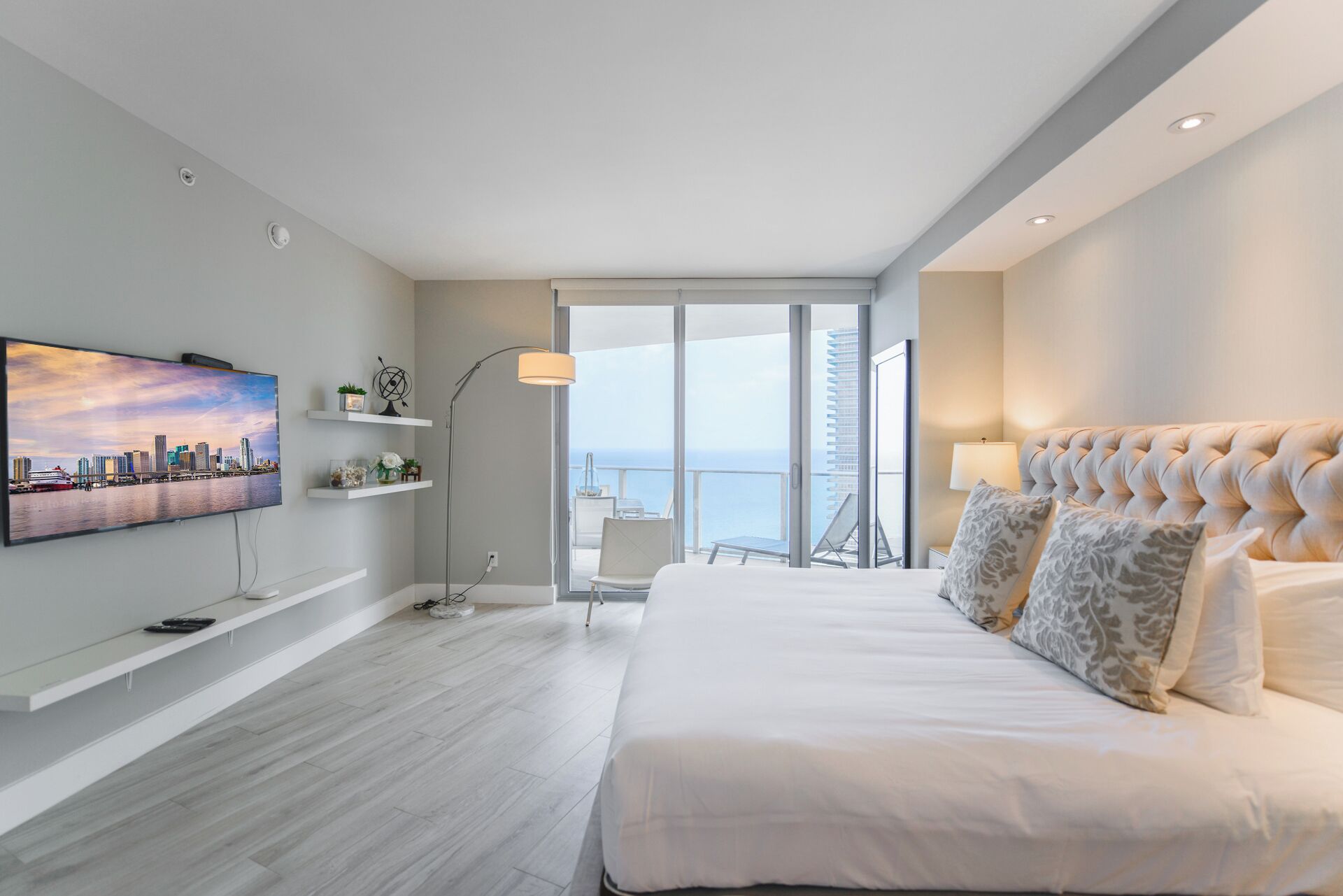 Une chambre d’hôtel moderne avec un grand lit, un tableau représentant un paysage urbain, un balcon avec vue sur la ville et une lampe sur pied.