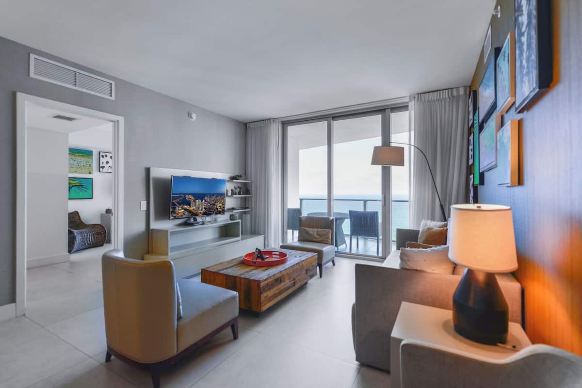 Un salon moderne doté d’un téléviseur à écran plat, de sièges confortables, d’une table basse en bois et d’un balcon offrant une vue sur la mer.