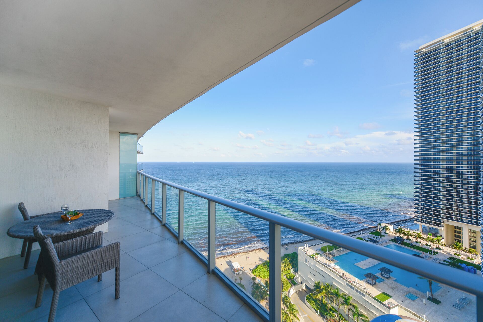 A balcony with a view of the ocean, a dining table, and a building with a pool.