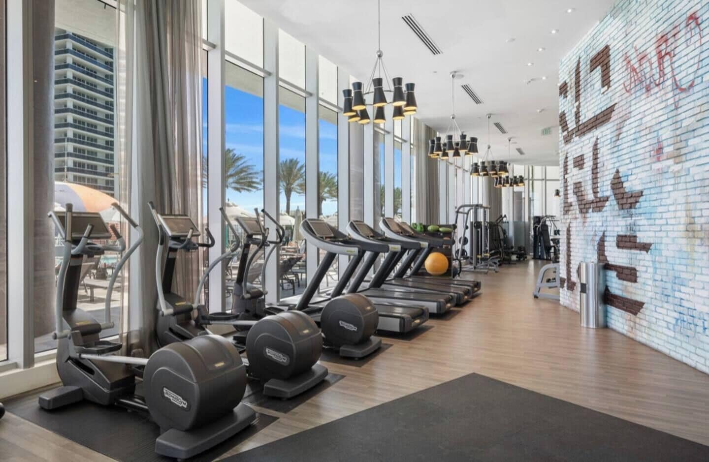 Een moderne sportschool met loopbanden, elliptische trainers en gewichten. Grote ramen bieden uitzicht op palmbomen en een gebouw buiten.