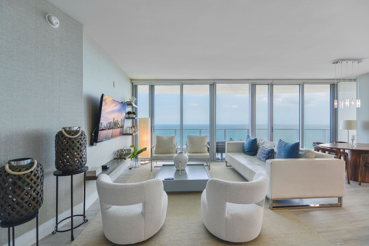 Un salon moderne doté d’une grande fenêtre donnant sur la mer, d’un téléviseur à écran plat et d’un coin salon confortable avec un canapé et des fauteuils.