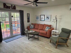 Sala de estar com sofá, poltrona, mesa de centro, ventilador de teto e porta que dá para um deck externo.