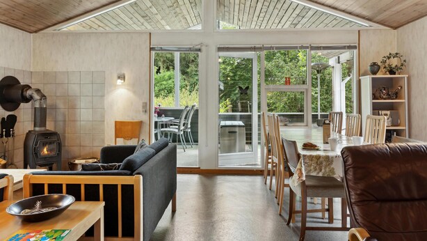 Een gezellig interieur met een houtkachel, een eethoek met stoelen en een tafel, en een groot raam met uitzicht op groen.