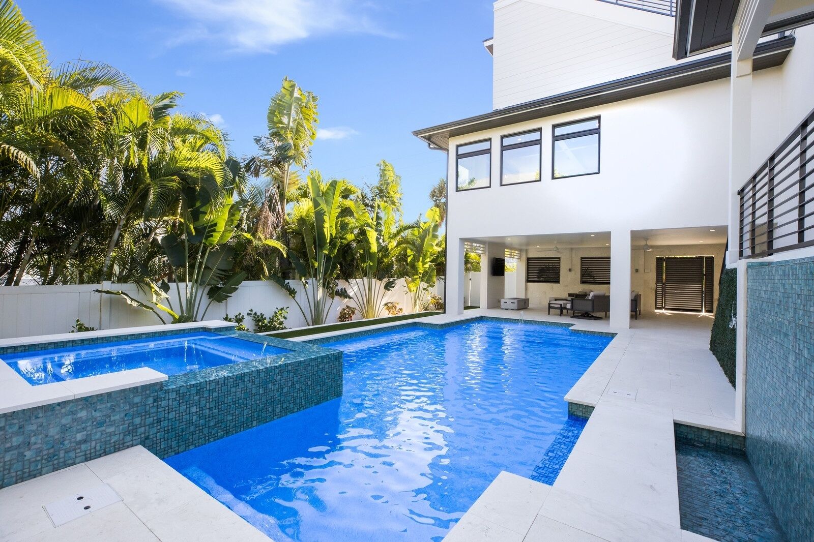 Uma casa moderna com piscina, um alpendre com mobiliário de exterior e plantas tropicais exuberantes.