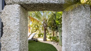 A stone archway leading to a garden with a lawn, palm trees, and a lounge chair. - Sizzling Summer Rates at Surf Side Beach House  GULF VIEWS Elevator (Beach Retreats) (Holmes Beach)