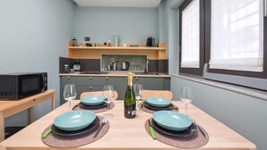 Une cuisine compacte équipée d’un four à micro-ondes, d’une machine à café et de divers ustensiles rangés sur des étagères. Une table est dressée avec des assiettes bleues, des verres à vin et une bouteille de champagne.