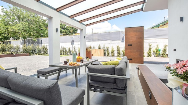 Une terrasse extérieure moderne avec un toit en verre, des meubles gris et une vue sur un immeuble et des arbres.