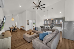A spacious living area with a large gray sofa, wooden dining table, and a kitchen with modern appliances.