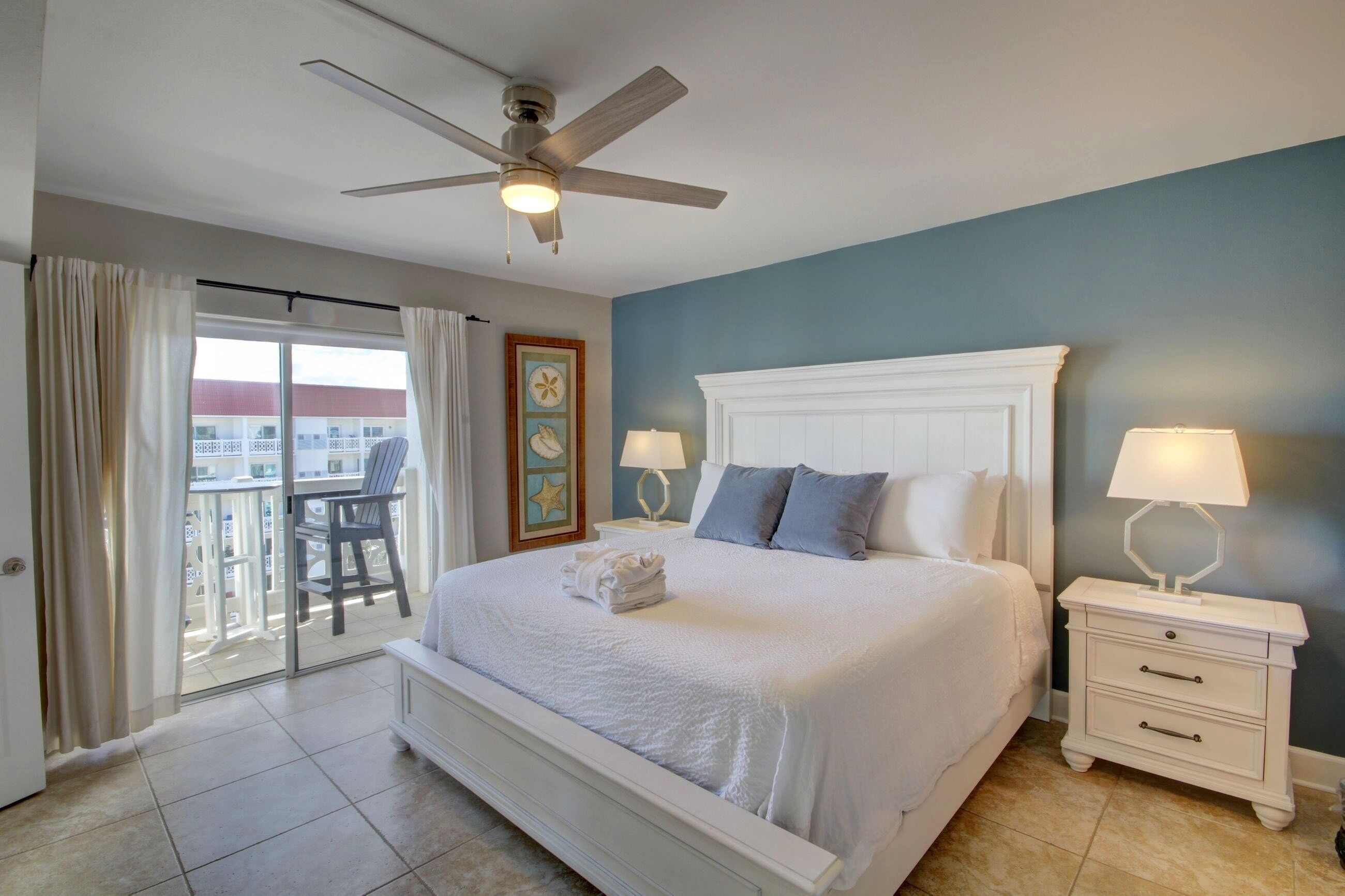 A bedroom with a bed, two bedside tables, a ceiling fan, a balcony with a chair, and a view of buildings outside.