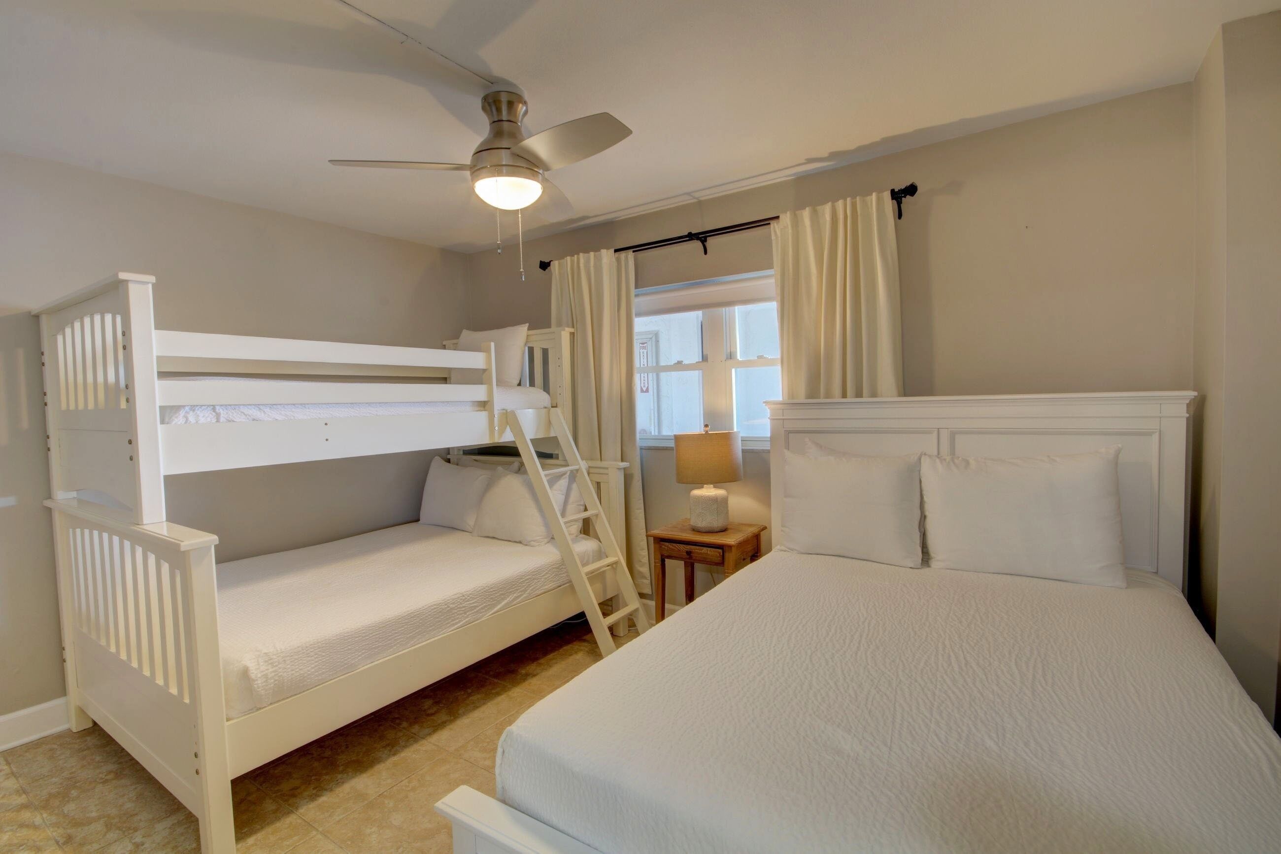 A bedroom with a bunk bed, a single bed, a ceiling fan, a window with curtains, and a bedside lamp.
