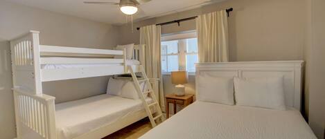A bedroom with a bunk bed, a single bed, a ceiling fan, a window with curtains, and a bedside lamp.