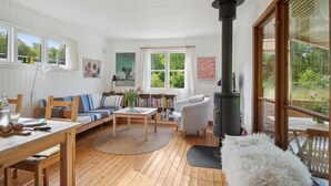 Ein gemütliches Wohnzimmer mit Holzboden, einer Couch, einem Couchtisch, einem Bücherregal, einem Kamin und großen Fenstern mit Blick auf viel Grün.