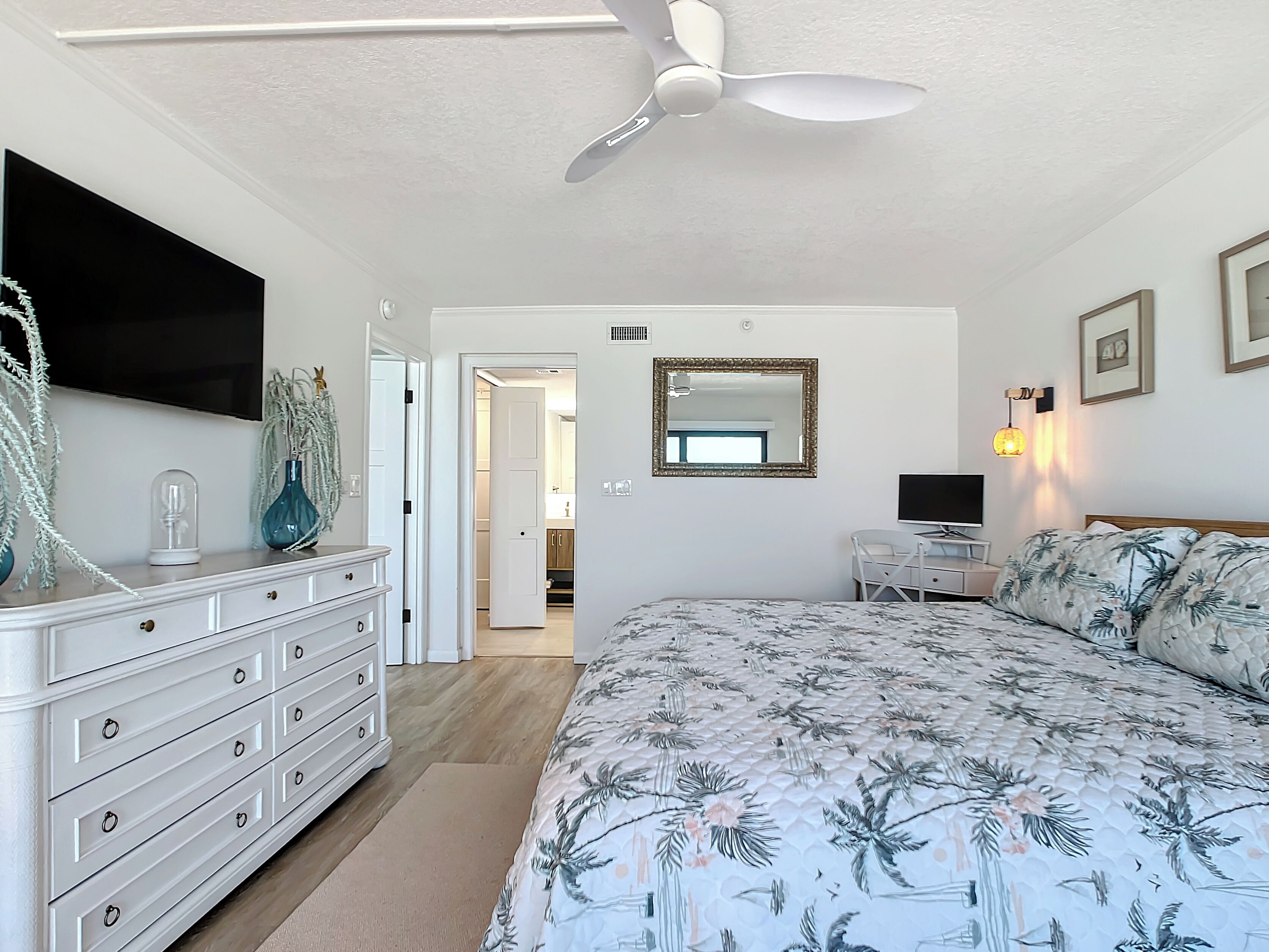 Un dormitorio con cama, cómoda, ventilador de techo, televisor y puerta que da a un baño.
