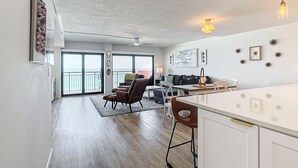 Un vaste espace de vie avec une grande porte-fenêtre coulissante donnant sur un balcon, un coin repas avec une table et des chaises blanches, et une cuisine dotée d’un comptoir blanc et de tabourets de bar.