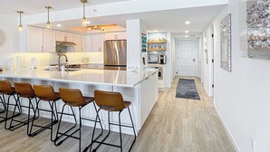 Une cuisine moderne dotée d’un îlot central, de tabourets de bar, d’appareils électroménagers en acier inoxydable, de luminaires suspendus et d’un couloir menant à une porte.