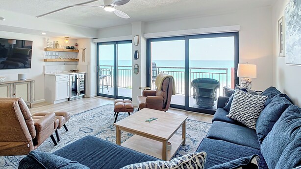 Un salon spacieux avec un grand canapé, une table basse et un ventilateur de plafond. Il y a un balcon avec vue sur la mer.