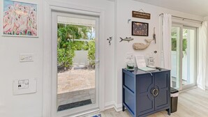 A modern interior with a large sliding glass door leading to a patio, a dark blue cabinet with a decorative item, and wall-mounted artwork including a flamingo painting and a sign. - Salt Air East managed by Beach Retreats (Holmes Beach)