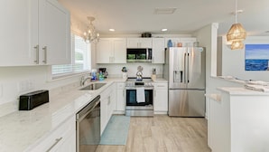 A modern kitchen with white cabinetry, stainless steel appliances, and a view of the ocean through the window. - Salt Air East managed by Beach Retreats (Holmes Beach)