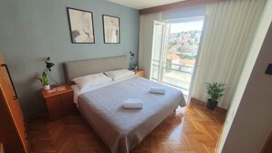 A bedroom with a large bed, two nightstands, a window with curtains, and a view of buildings outside. - Apartment Carrington (Dubrovnik)