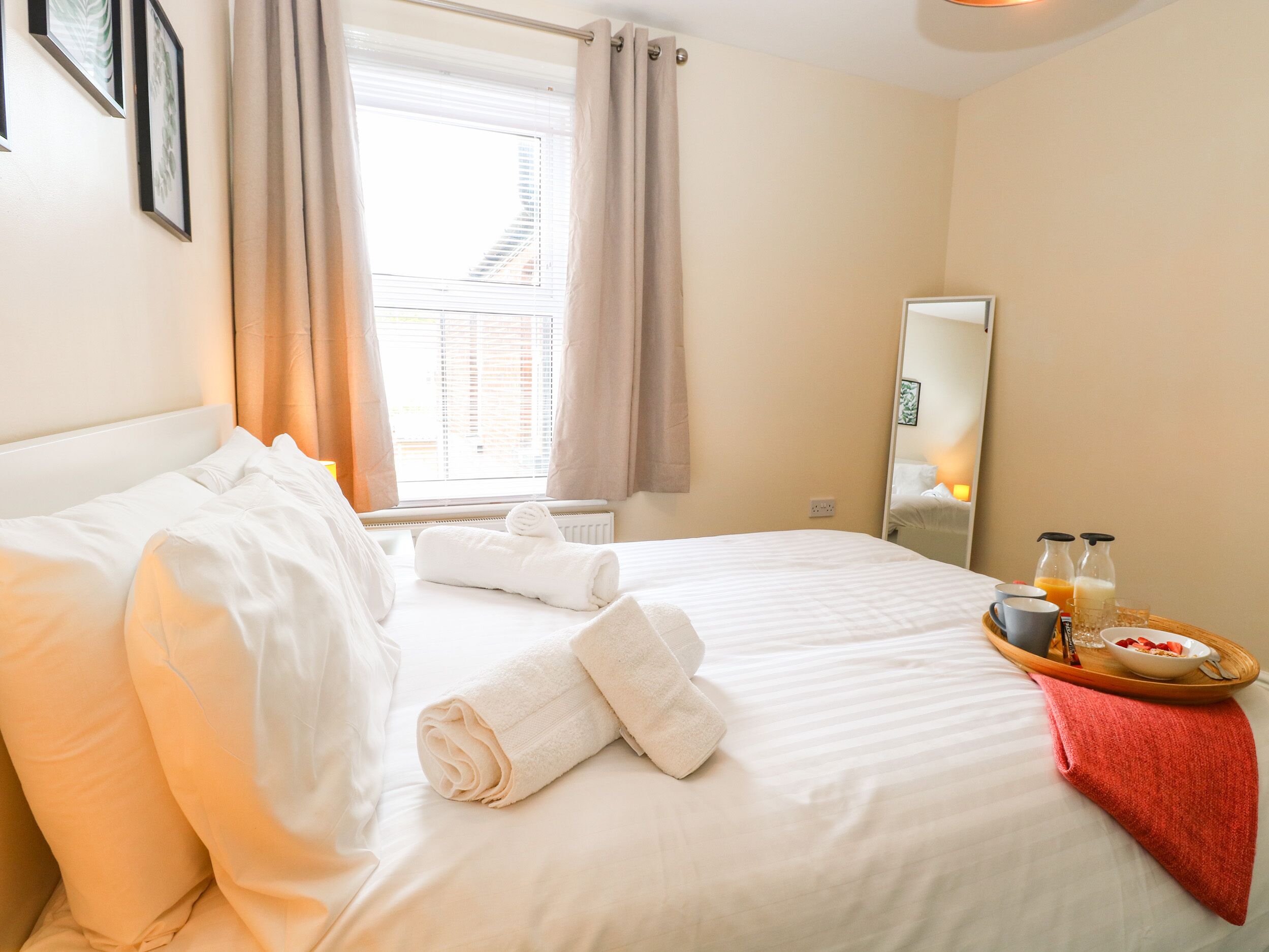 Una habitación de hotel con una cama bien hecha, una ventana con cortinas, una mesita de noche con una bandeja de desayuno y un espejo en la pared.