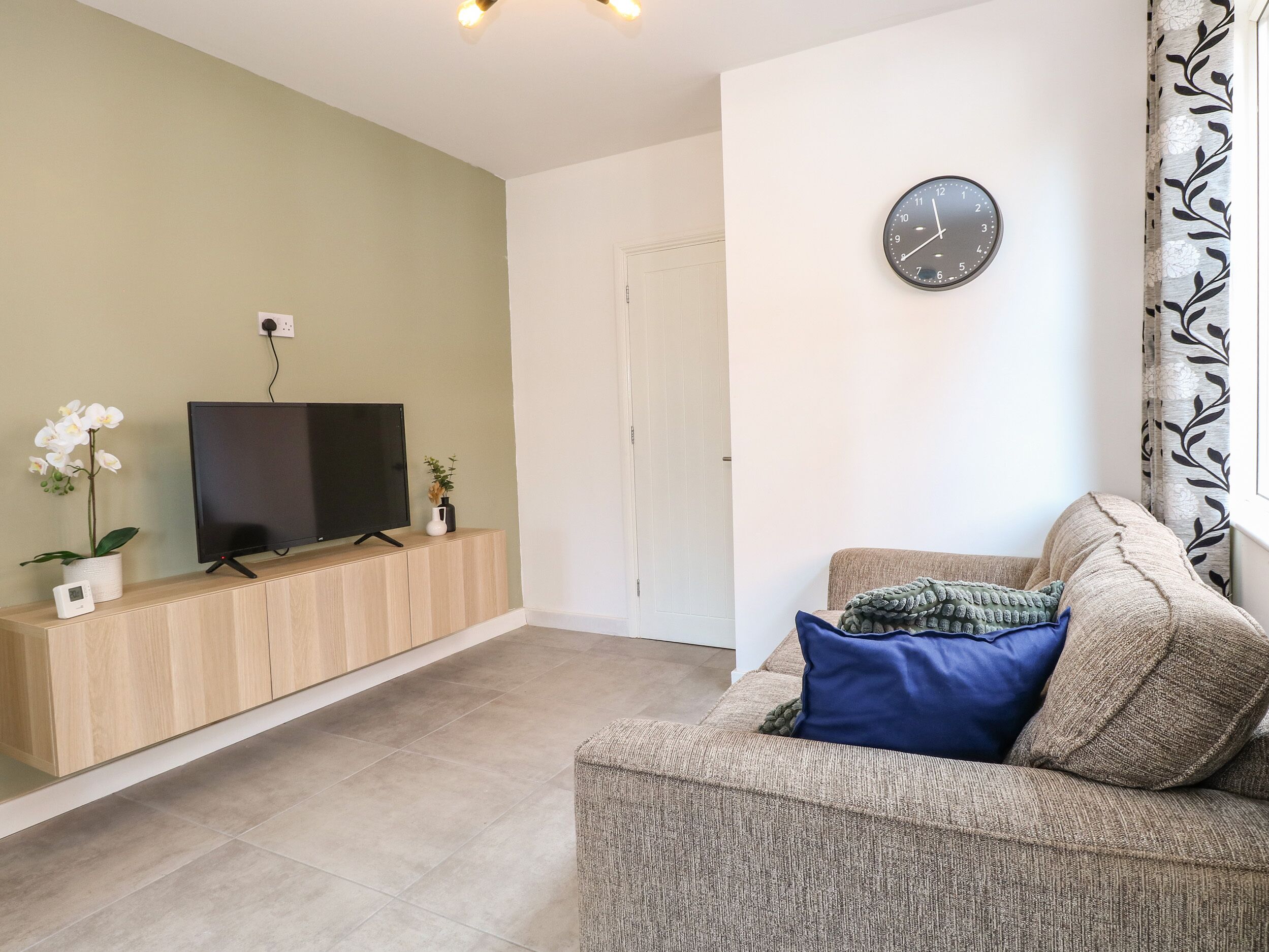 A living room with a TV on a wooden cabinet, a clock on the wall, and a sofa with cushions.