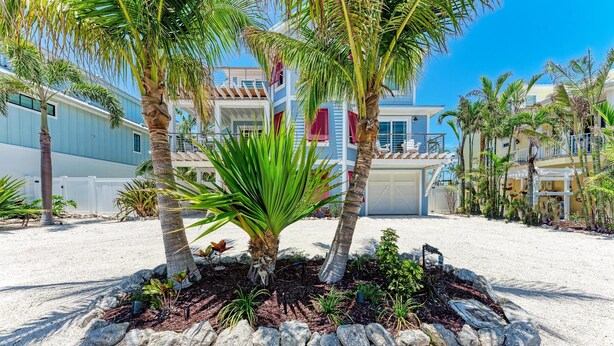 A beachfront property with palm trees, a sandy driveway, and a large building with a blue facade and red accents. - Chasing Sunsets managed by Beach Retreats (Holmes Beach)