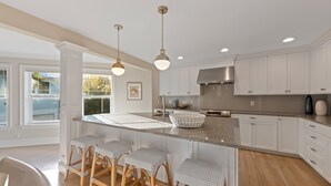 A spacious kitchen with a large island, white cabinetry, and a stainless steel range hood. There are three bar stools at the island and a window providing natural light. - Chasing Sunsets managed by Beach Retreats (Holmes Beach)