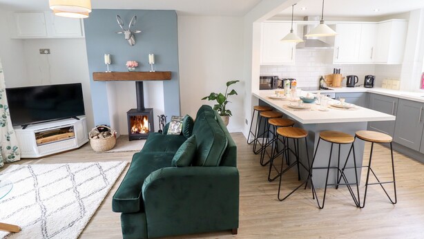 A modern living room with a green sofa, a flat-screen TV, a fireplace, and a kitchen with bar stools. - 6 Hillside Cottages (Gainsborough)
