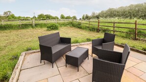 An outdoor patio with wicker furniture and a stone floor, surrounded by a grassy area and a wooden fence. - 6 Hillside Cottages (Gainsborough)