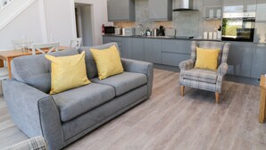 A modern living room with a grey sofa and armchair, both with yellow cushions, and a wooden dining table with chairs. A staircase is visible in the background. - Willow Cottage (Ilfracombe)