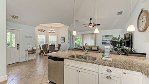 A spacious kitchen with granite countertops, white cabinetry, and a dishwasher. There is a dining area with wicker furniture and a view of the outdoors. - Starfish Isle managed by Beach Retreats (Holmes Beach)