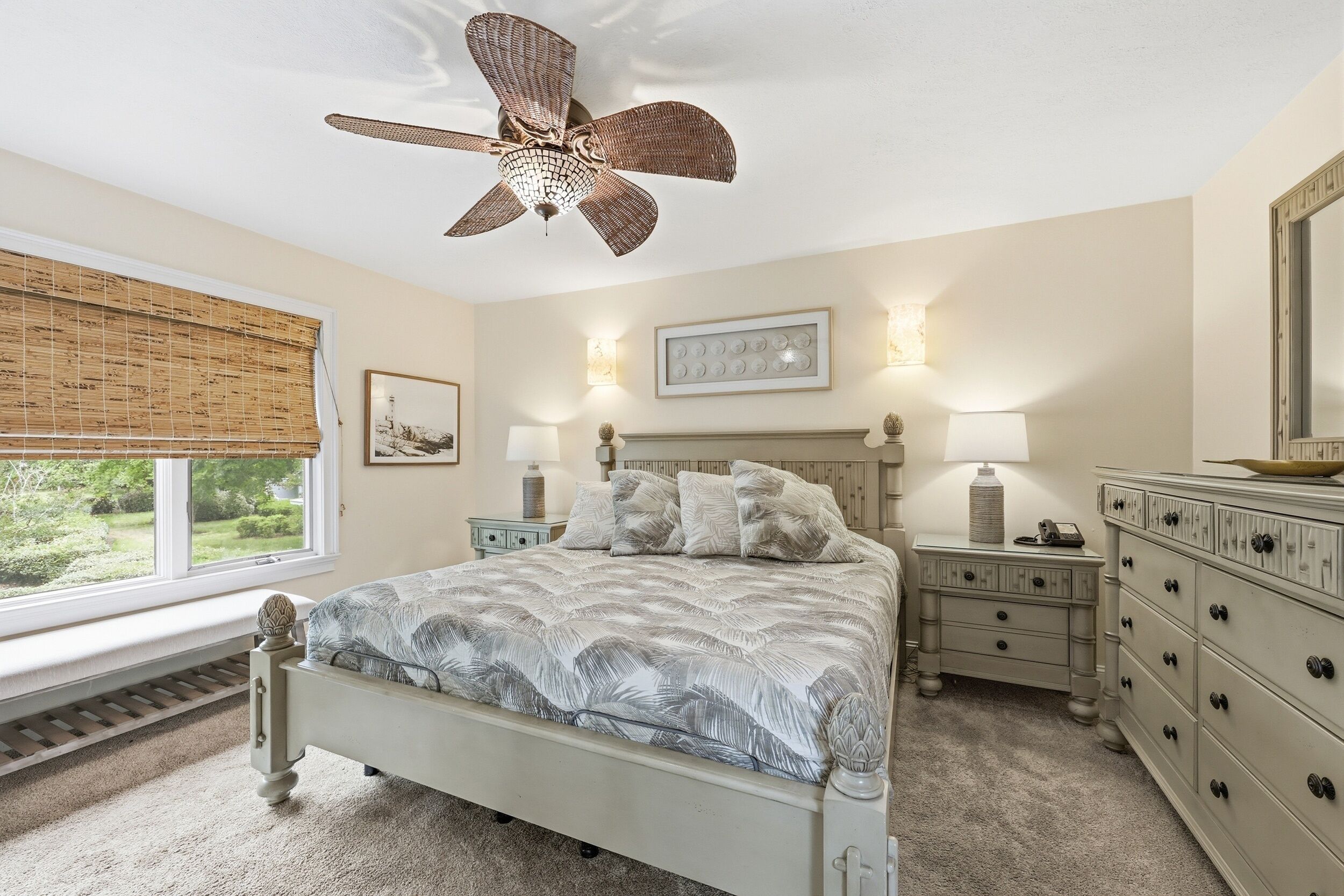 A bedroom with a bed, nightstands, dresser, ceiling fan, window with blinds, and wall-mounted lamps.