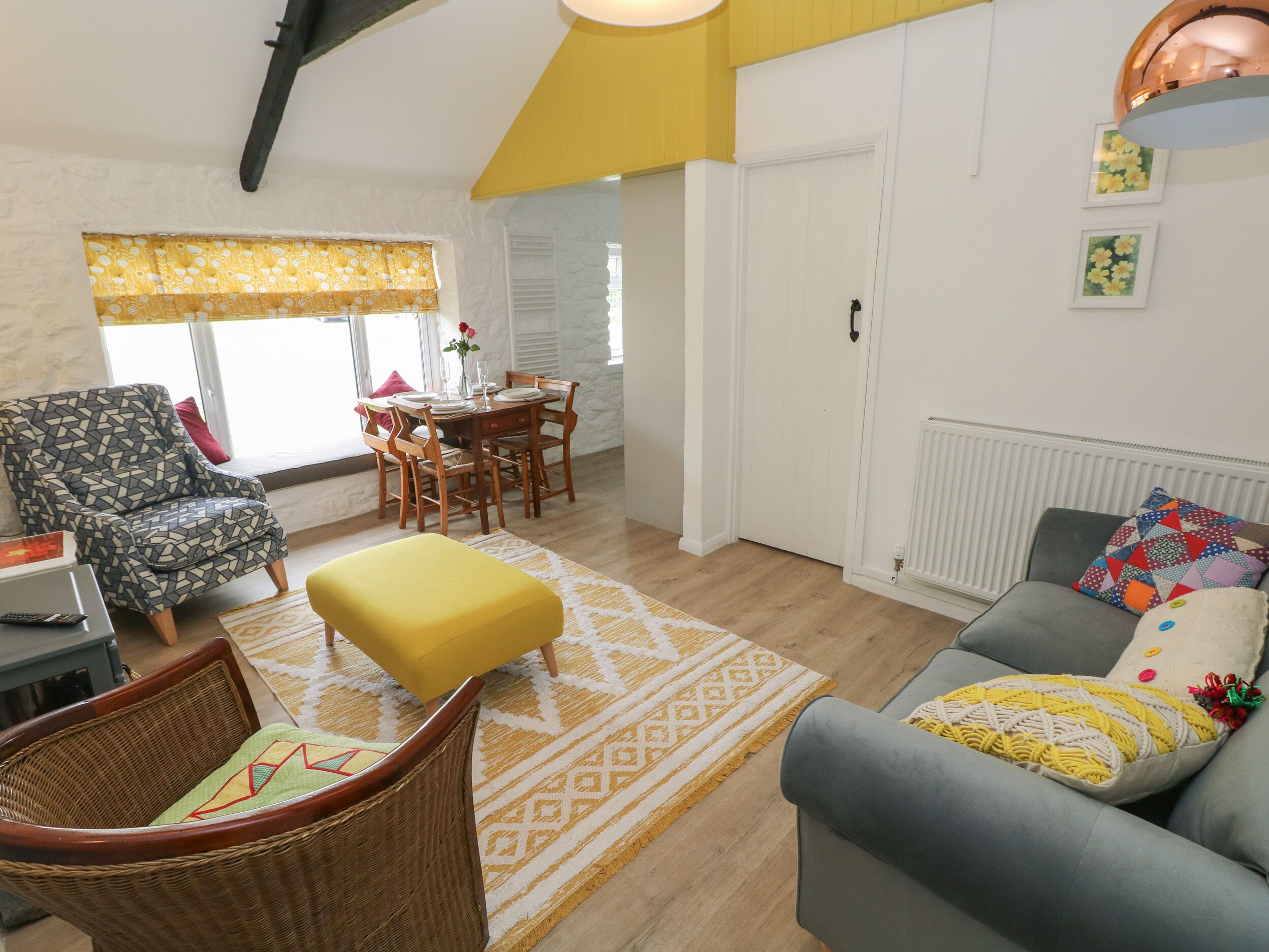Un salon comprenant un coin repas, un canapé gris, un pouf jaune, un fauteuil en osier et un tapis à motifs.