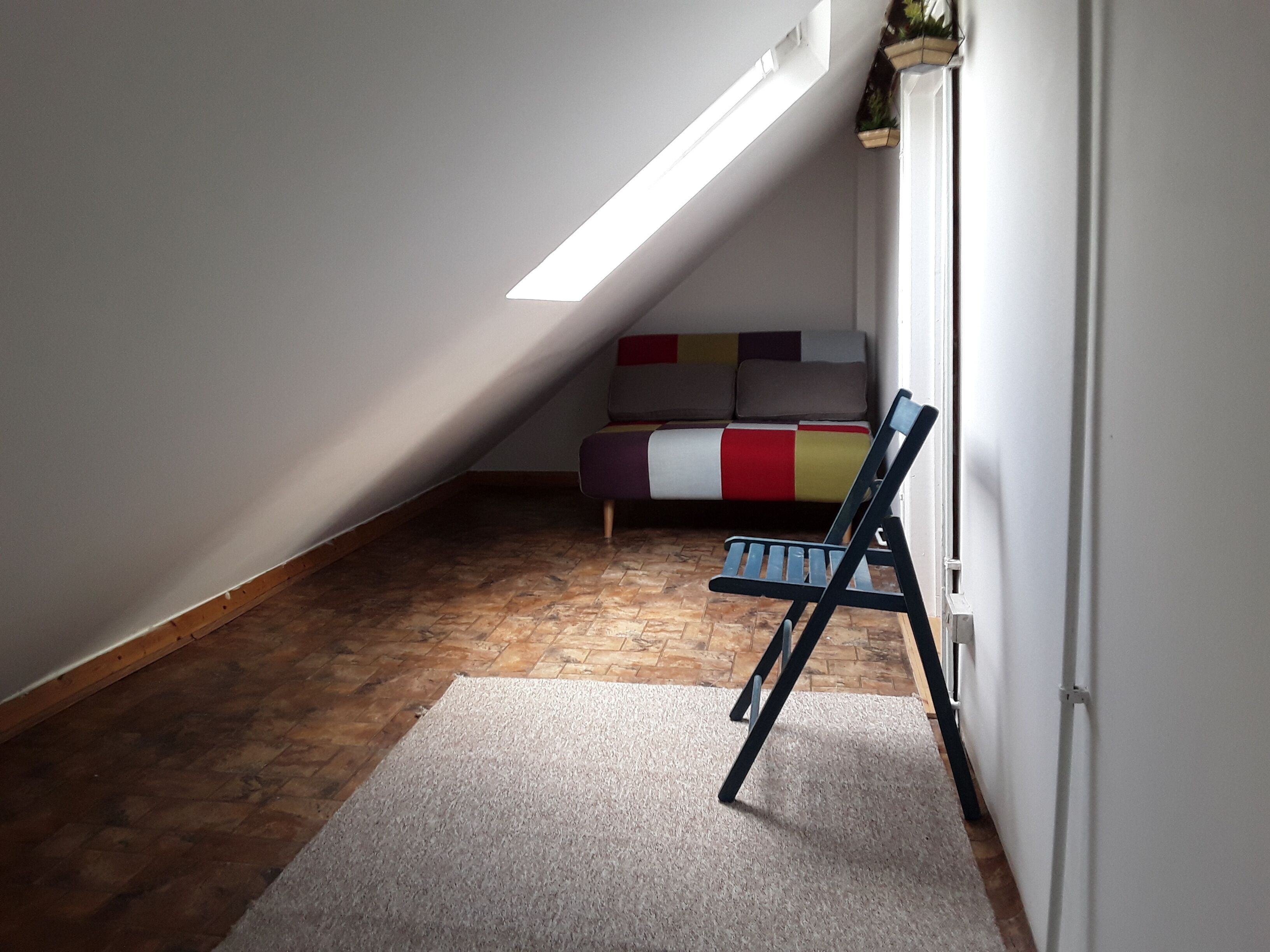 Une petite pièce avec un plafond en pente, un plancher en bois, un canapé coloré et une chaise bleue.