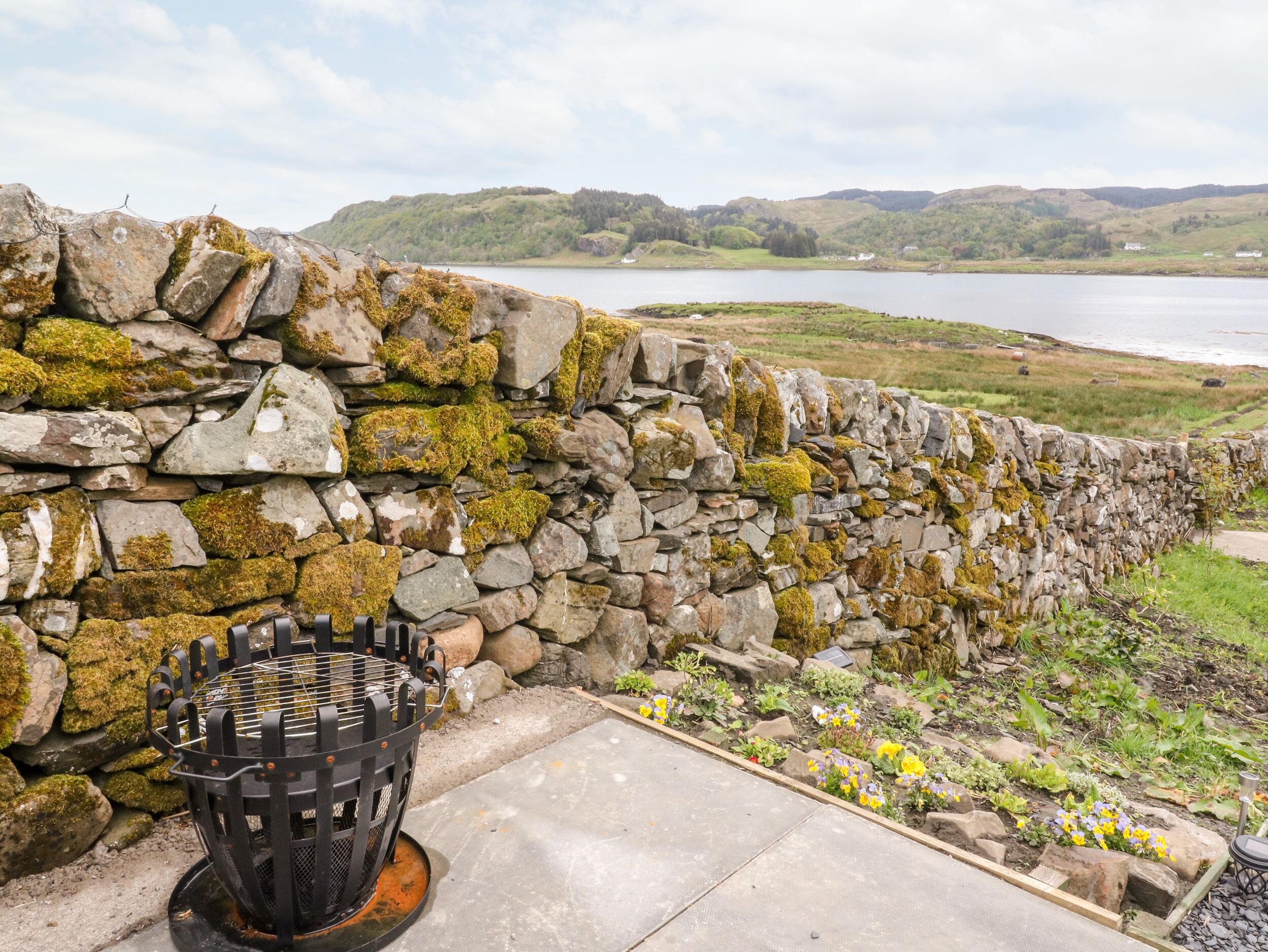 A stone wall with a metal grill, a concrete pathway, and a flower bed with yellow and purple flowers.