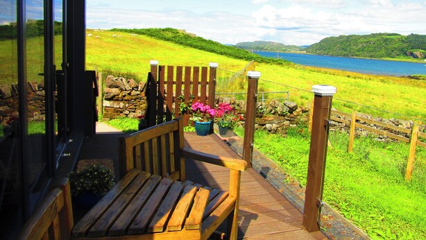 Eine Holzterrasse mit einer Bank, die auf ein Wiesenfeld und ein Gewässer blickt.