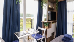 Ein Zimmer mit einem Bett, einem Schreibtisch und einem Stuhl. Ein Fenster mit Vorhängen, ein Fernseher auf einem Regal und eine Pflanze auf dem Regal.