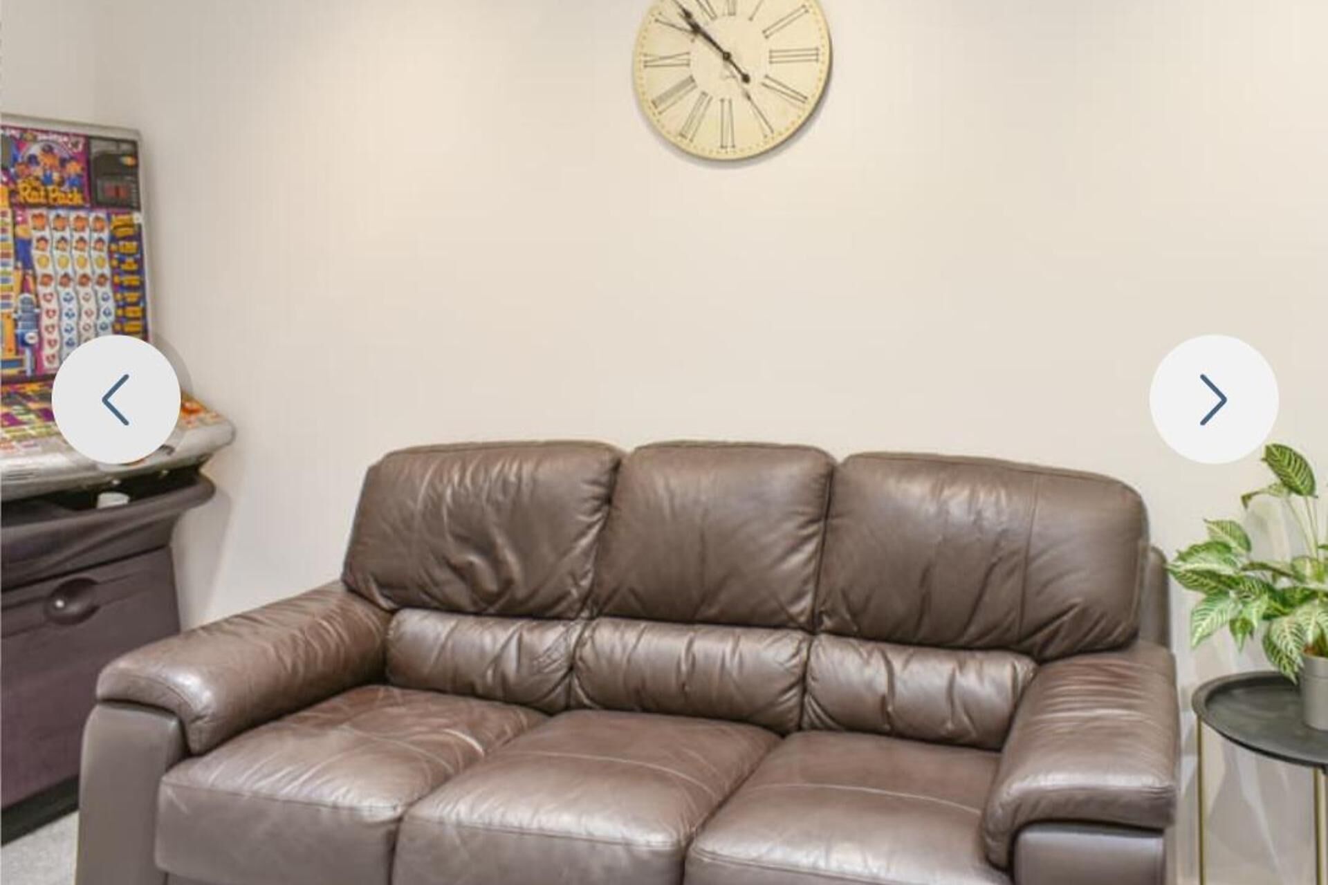 Ein braunes Ledersofa in einem Raum mit einer Uhr an der Wand und einem kleinen Tisch mit einer Pflanze.