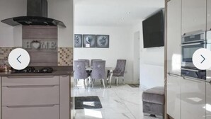 A modern kitchen with a built-in stove and a range hood, a dining area with a table and chairs, and a living area with a sofa and a TV. - The Hawthorns Large Detached 3 Bedroom Family Home (Seaham)
