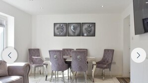A dining area with a white table and six chairs, a sofa, and three framed artworks on the wall. - The Hawthorns Large Detached 3 Bedroom Family Home (Seaham)