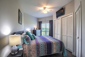 A bedroom with a bed, a ceiling fan, a TV mounted on the wall, and a window with curtains.